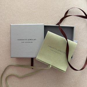 May include: A light gray square jewelry box with the text Serenity Jewelry Los Angeles printed in black. Inside, a sage green fabric pouch features the same text. A dark brown ribbon is draped across the open box and the green pouch.