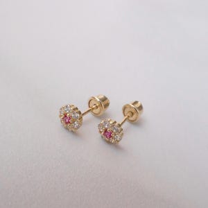 May include: A pair of gold stud earrings. Each earring has a flower-shaped design with a central pink gemstone surrounded by white gemstones. The earrings have a screw-back fastening.