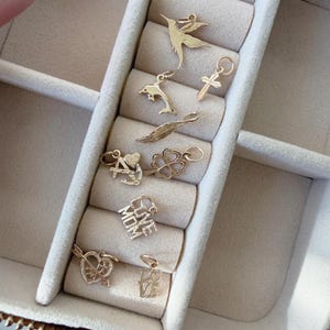 May include: A collection of gold-toned metal charms including a hummingbird, a dolphin, a cross, a feather, an anchor, a four-leaf clover, a heart with an arrow, and two charms with the text LOVE MOM and LOVE, each featuring a small jump ring.