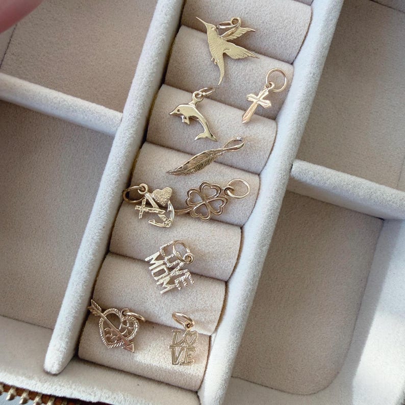 May include: A collection of gold-toned metal charms including a hummingbird, a dolphin, a cross, a feather, an anchor, a four-leaf clover, a heart with an arrow, and two charms with the text LOVE MOM and LOVE, each featuring a small jump ring.