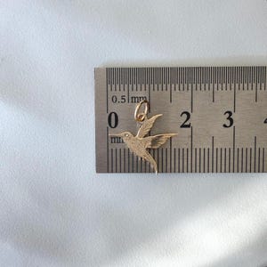 May include: A gold-toned hummingbird pendant charm with textured wing and body details, positioned against a metal ruler showing a width of approximately 0.75 inches from the beak to the tail tip and a height of about 0.75 inches.