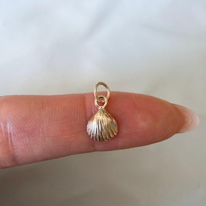 May include: A small, gold-toned metal charm shaped like a seashell, featuring a ribbed texture on its surface and a circular jump ring attached to the top for hanging. The charm has a polished finish and a distinct fan-like scallop shell design.
