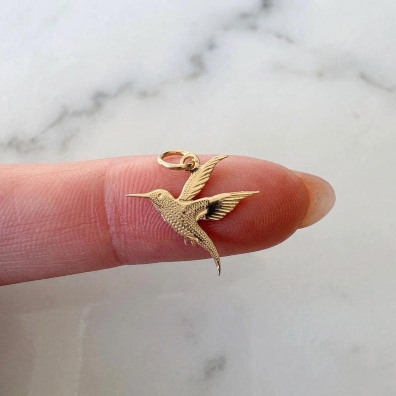 May include: A small gold-toned metal charm shaped like a hummingbird in flight. The charm features detailed textured wings and a pointed beak, with a circular jump ring attached at the top for hanging. The design captures the bird in a mid-air pose.