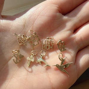 May include: A collection of eight small gold charms featuring various designs including a heart with an arrow, a four-leaf clover, the text LOVE MOM, the word LOVE, an anchor with a heart, a cross, a dolphin, and a hummingbird in flight.
