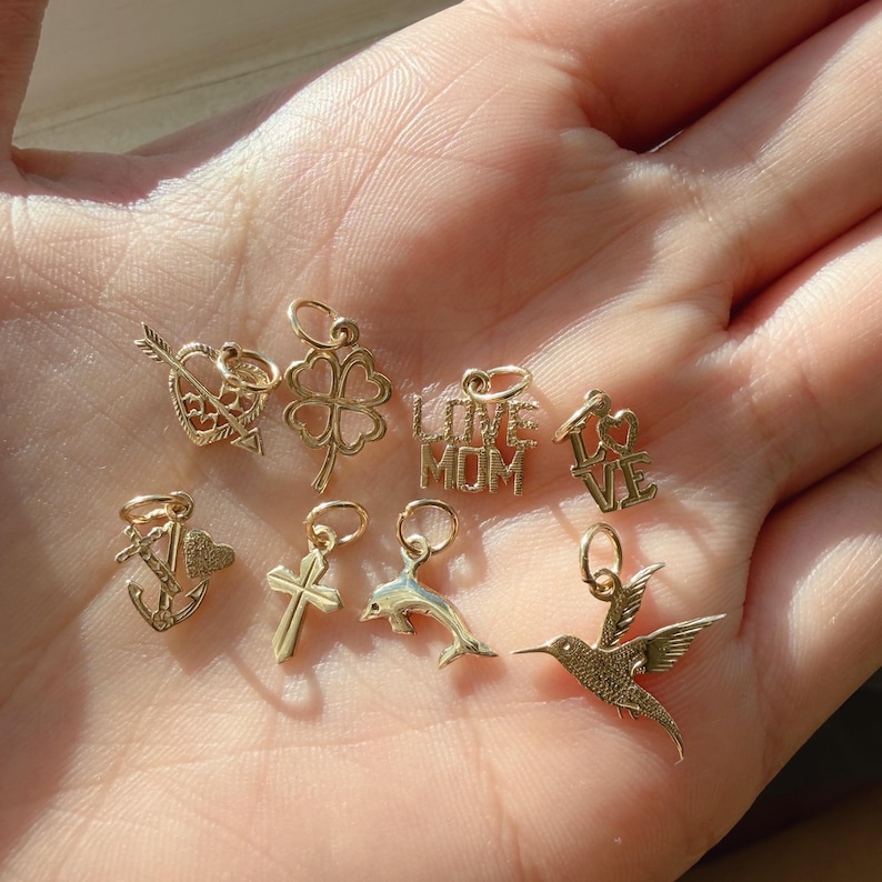 May include: A collection of eight small gold charms featuring various designs including a heart with an arrow, a four-leaf clover, the text LOVE MOM, the word LOVE, an anchor with a heart, a cross, a dolphin, and a hummingbird in flight.