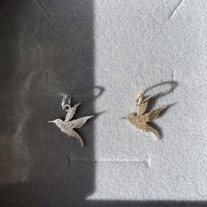 May include: Two hummingbird pendants, one in a silver-toned metal and one in a gold-toned metal. Each pendant features a detailed, textured bird in flight with wings extended and a small jump ring attached to the top for hanging on a chain.
