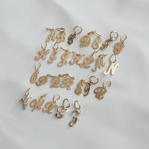 May include: A collection of gold-colored initial charms, each with a loop for attachment. The charms are in a cursive font and are arranged on a white surface. The letters A, B, C, D, E, F, G, H, I, J, K, L, M, N, O, P, Q, R, S, T, V, W, X, Y, and Z are visible.