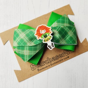 May include: A green hair bow with a plaid pattern and a cartoon image of a red-haired girl in a green dress. The bow is on a brown cardboard backing with the text "Starchasers Handmade in Scotland with love".