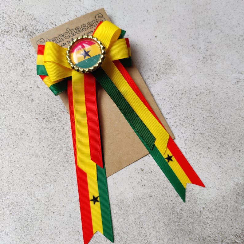 May include: A yellow, red, and green ribbon bow with a gold button featuring the flag of Ghana. The bow is attached to a brown card with the text "Starchasers" printed on it.