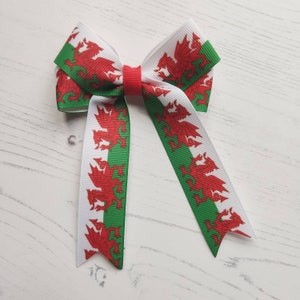 May include: A white bow with red and green ribbon featuring the Welsh dragon. The bow is made of fabric and has a white background with red dragons and green stripes.