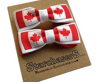 May include: Two red and white Canadian flag hair bows with a white background. The bows are on a brown card with the text "Starchasers Handmade in Scotland with love .co.uk"