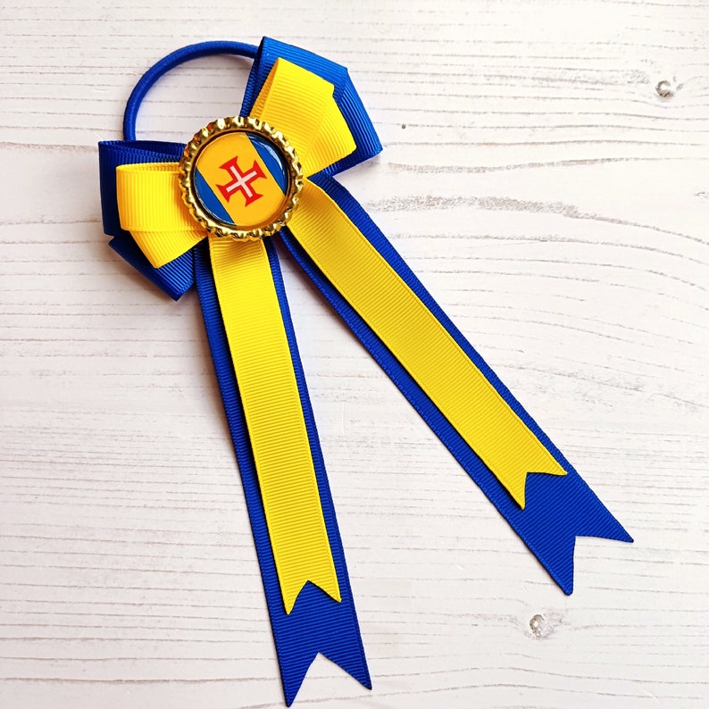 May include: A yellow and blue hair bow with a gold button featuring the flag of the Azores. The bow is tied with a blue ribbon.