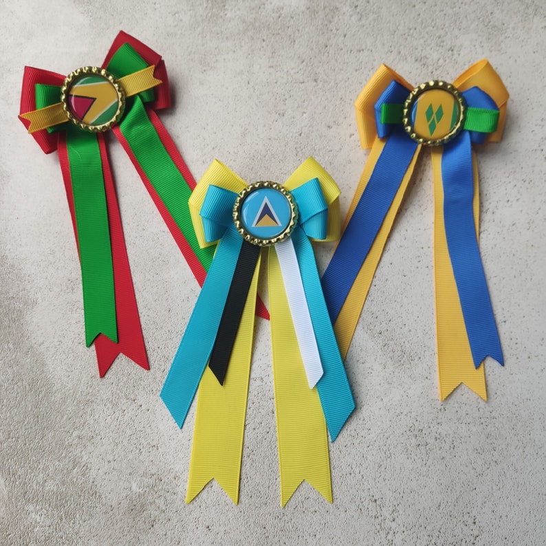 May include: Three handmade hair bows with different colored ribbons and a small button with a country flag design in the center. The bows are yellow, blue, and green with red, white, and black ribbons.