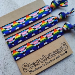 May include: Three rainbow-patterned hair ties featuring transgender heart symbols, displayed on a brown card. The card is printed with the text "Starchasers" and "Handmade in Scotland with love."