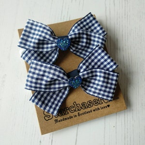 May include: Two blue and white gingham hair bows with blue glitter heart centres. The bows are on a brown card with the text "Sarchaser Handmade in Scotland with love".