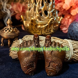 May include: Two bronze-colored foot-shaped amulets featuring embossed fish designs on the soles and small spider motifs on the toes. The text etsy.com/shop/ArabicSpells is printed in green across the center of the two objects.