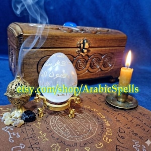 May include: A smooth, white, egg-shaped stone featuring gold Arabic calligraphy, resting on a gold-toned metal stand. Beside it sits a gold-toned metal incense burner, a lit yellow candle, and a wooden chest with intricate carvings.