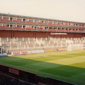 Puede incluir: Una vista de un estadio de fútbol con una pared de ladrillo y un gran campo de césped. El estadio tiene una zona de asientos de varios niveles con asientos rojos y un gran marcador con el texto "BARCLAYS BANK" visible. Hay varios anuncios en la pared y alrededor del campo.