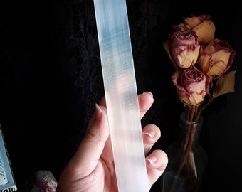 May include: A white selenite wand, also known as a cleansing wand, is held in a hand. The wand is smooth and has a rectangular shape.
