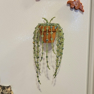 May include: A decorative hanging plant ornament featuring a small terracotta-colored pot filled with cascading strands of light green and iridescent glass beads that mimic the appearance of a trailing succulent plant.