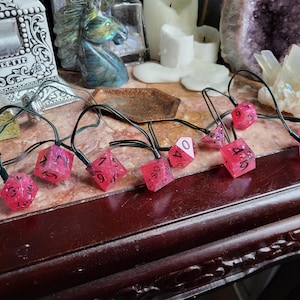 May include: A set of eight pink and white dice with black numbers, strung together with black wire and small light bulbs. The dice are on a dark wooden surface.