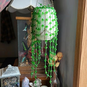 May include: A hanging plant decoration featuring a white ceramic pot and long, cascading strands of bright green faceted beads. The plant is suspended by a thin gold-toned metal chain, creating a trailing effect that mimics a string of pearls.