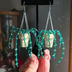 May include: A pair of dangling earrings featuring miniature cream-colored pots holding beaded green trailing plants. The plants are constructed from shimmering green beads on thin wires, suspended by silver-toned metal chains from earring hooks.