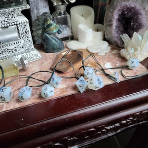 May include: A set of ten iridescent white dice with black numbers, strung together with black cord. The dice are arranged in a semi-circle on a dark brown surface.