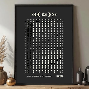 May include: A framed black and white 2026 moon phase calendar. The calendar displays the moon phases for each day of the year, with the city of New York at the bottom. The calendar is surrounded by decorative vases and a plant.