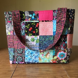 Peut inclure: Un sac fourre-tout en patchwork avec des carr&eacute;s de tissu noir, rose, bleu et vert. Le sac a deux anses noires.