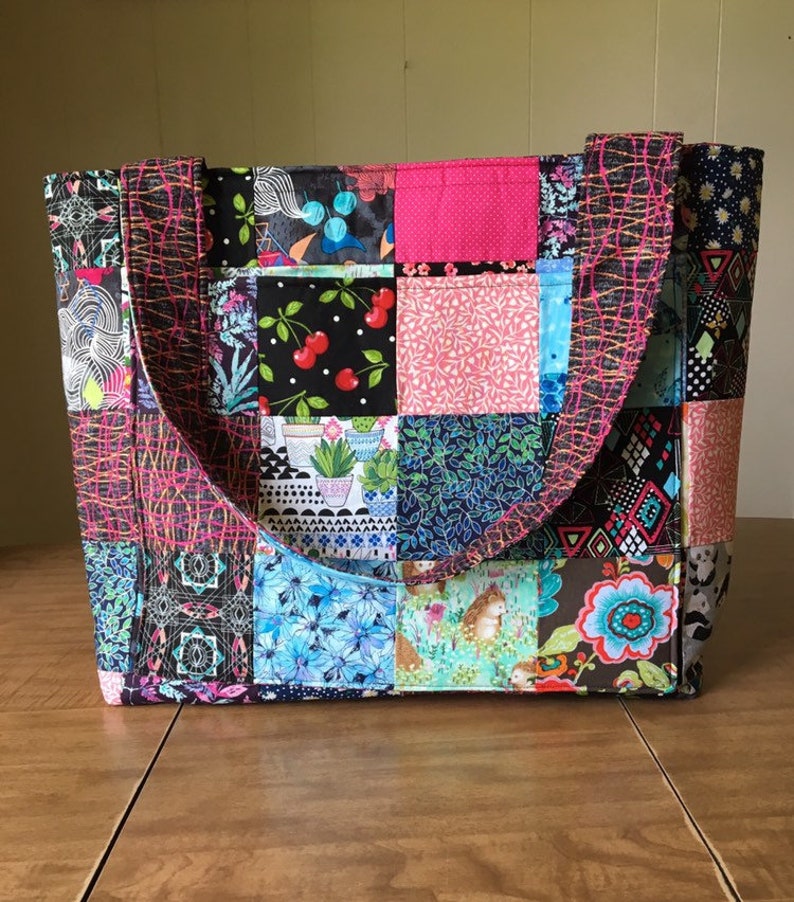 Peut inclure: Un sac fourre-tout en patchwork avec des carr&eacute;s de tissu noir, rose, bleu et vert. Le sac a deux anses noires.