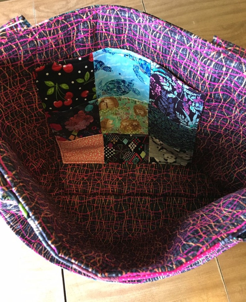 Peut inclure: L'int&eacute;rieur d'un sac fourre-tout noir et rose avec une poche en patchwork faite de diff&eacute;rents tissus color&eacute;s. La poche est compos&eacute;e de 9 carr&eacute;s de tissu avec diff&eacute;rents motifs, dont des cerises, des tortues et des fleurs.