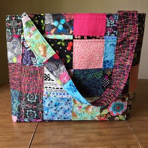 Peut inclure: Un sac fourre-tout en patchwork avec une vari&eacute;t&eacute; de tissus color&eacute;s, dont du rose, du bleu, du vert et du noir. Le sac a deux longues anses et un motif en patchwork.