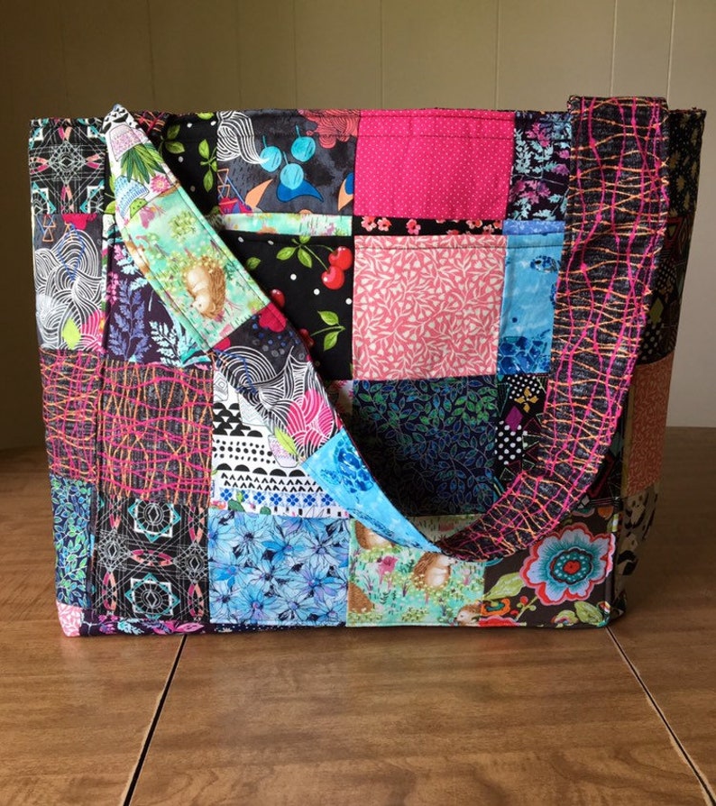 Peut inclure: Un sac fourre-tout en patchwork avec une vari&eacute;t&eacute; de tissus color&eacute;s, dont du rose, du bleu, du vert et du noir. Le sac a deux longues anses et un motif en patchwork.