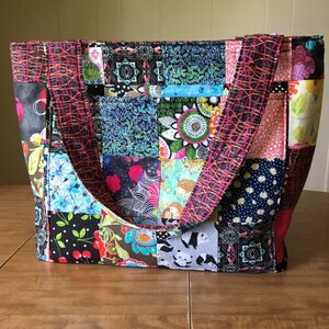 Peut inclure: Un sac fourre-tout en patchwork avec des carr&eacute;s de tissu noir, blanc, rose, bleu et vert. Le sac a deux anses rose et noir.