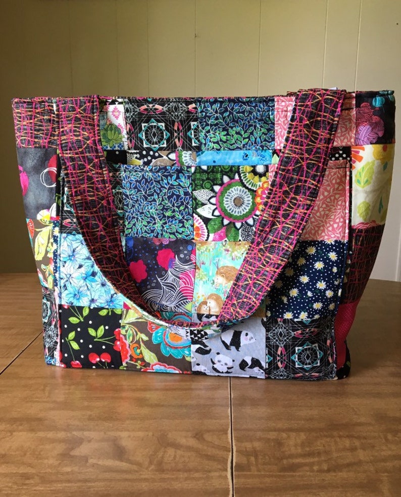Peut inclure: Un sac fourre-tout en patchwork avec des carr&eacute;s de tissu noir, blanc, rose, bleu et vert. Le sac a deux anses rose et noir.
