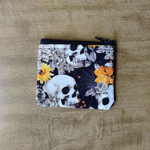 May include: A small, square coin purse with a black zipper. The fabric features a repeating pattern of white skulls, gray roses, yellow and orange flowers, and a spiderweb design on a black background.