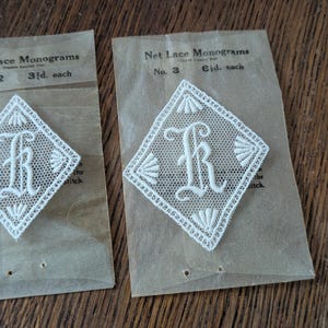 May include: Two vintage white net lace monograms, each with the letter "R" in a diamond shape. They are presented on aged paper packaging with the text "Net Lace Monograms". The background is a wooden surface.