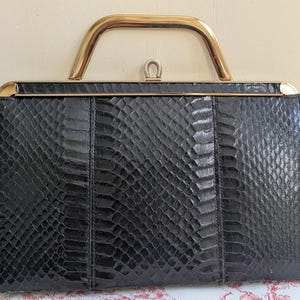 May include: A black snakeskin-patterned handbag with a gold-coloured handle. The rectangular bag has a structured design with a clasp closure and gold trim. The handle is a curved, arched shape.