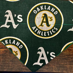 Op de afbeelding: Groen stof met een herhalend patroon van witte "A's" logo's. Sommige logo's zijn omgeven door een gele cirkel met de tekst "Oakland Athletics".