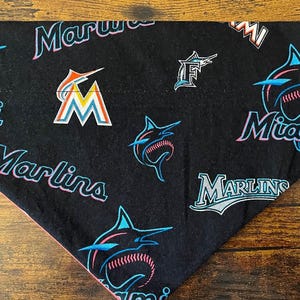 Op de afbeelding: Zwarte bandana met het Miami Marlins logo en teamnaam in teal, oranje en roze. Het ontwerp bevat het Marlins logo en het woord "Miami" in een gestileerd lettertype. De bandana heeft een roze rand.