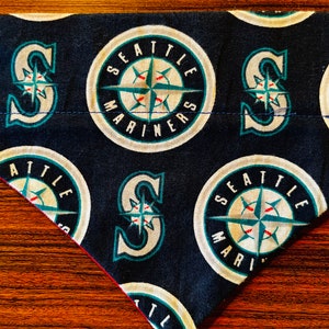 Può includere: Un bandana blu e bianco con un motivo che ripete il logo dei Seattle Mariners. Il logo presenta una bussola con una 'S' bianca e le parole 'Seattle Mariners' intorno alla bussola.