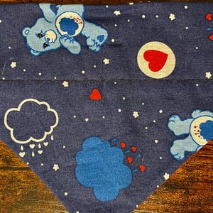 Pode incluir: Bandana azul escuro com tema Ursinhos Carinhosos, com um padrão repetido de Ursinhos Carinhosos, nuvens, corações e estrelas. O tecido tem detalhes em branco e azul claro. A bandana tem formato triangular.