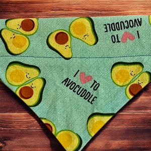 May include: A triangular pet bandana with a light teal background. The bandana features illustrations of avocados and the text "I love to avocuddle" in black. The heart is pink. The avocados are green, yellow, and brown.