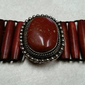 May include: A close-up of a necklace featuring a large, oval, reddish-brown stone set in a silver-colored metal frame. The necklace has cylindrical, reddish-brown beads and a black cord.