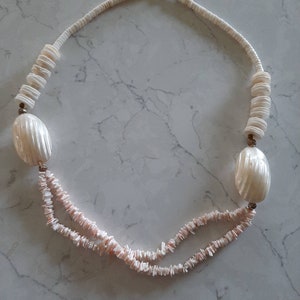 May include: A white shell necklace with two strands of small, irregular shell pieces. The necklace features two large, oval-shaped shell pendants and sections of stacked, round shell beads. The clasp is a dark metal.