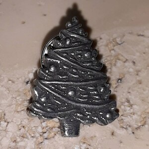 May include: A silver-colored Christmas tree pin. The tree has a detailed, textured design with small, round embellishments. The pin is a festive accessory, perfect for holiday celebrations or adding a touch of seasonal flair to clothing or accessories.
