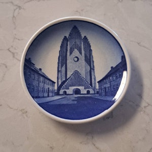 May include: A small, round, white and blue decorative plate featuring a detailed illustration of a tall church with a unique architectural design. The plate has a white border and a glossy finish.
