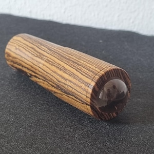 May include: A cylindrical bottle stopper made of wood with a dark brown and tan striped pattern. The stopper has a clear, rounded glass top. The product is designed to seal bottles.