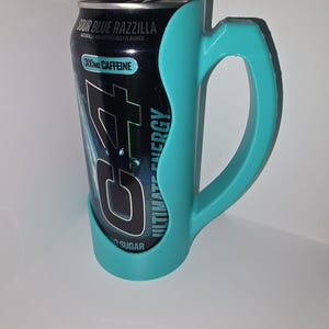 May include: A black energy drink can with the text "Sour Blue Razzilla" and "300mg Caffeine" is held by a teal-colored handle. The can is labeled "Ultimate Energy" and "No Sugar".